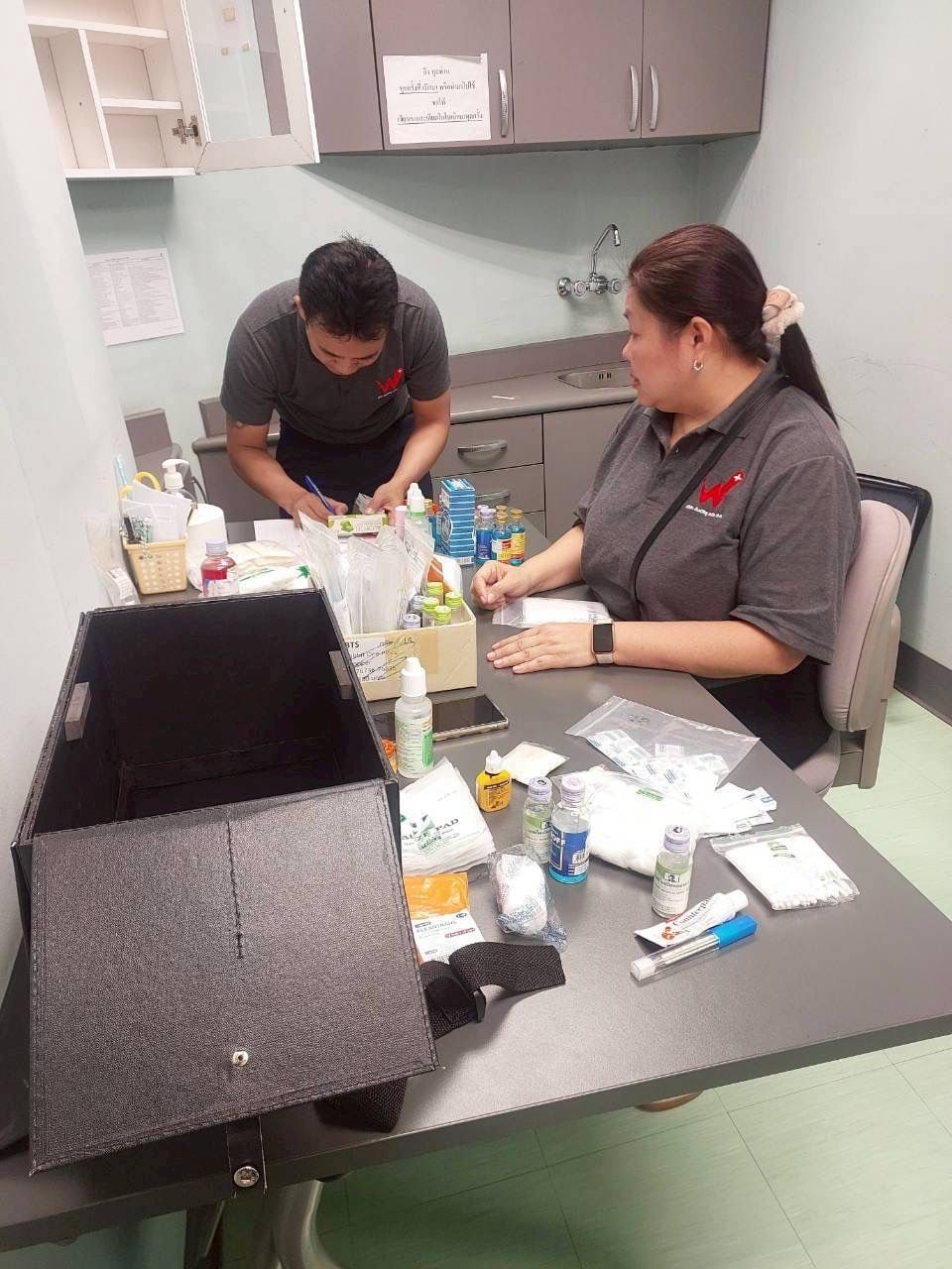 Two W-Asia staff in branded polos checking medical supplies at a first-aid room table
